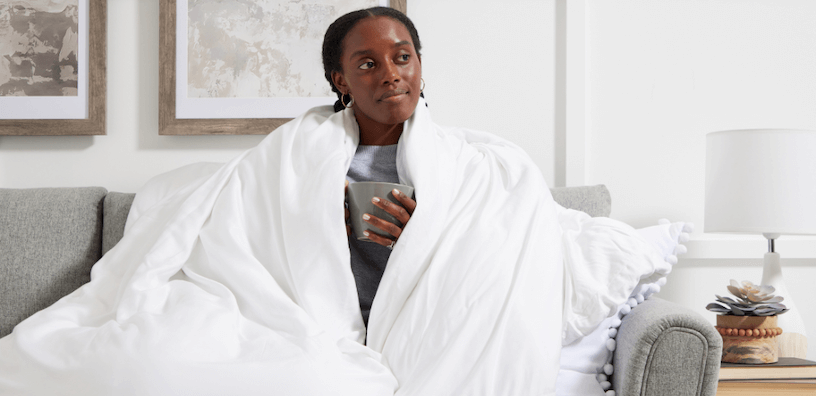 Eine Frau auf einem bequemen Sofa, eingehüllt in eine weiche weiße Decke, mit einer Tasse in der Hand und einem entspannten Gesichtsausdruck; eine ruhige und beruhigende Atmosphäre.