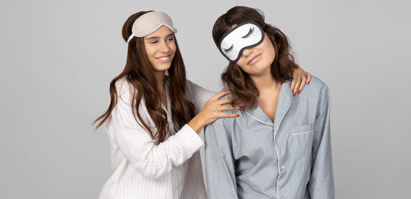 Zwei Frauen in Pyjamas und Schlafmasken. Die eine wirkt fröhlich und wach, die andere schläfrig, sie wird getröstet; symbolisiert die unterschiedliche morgendliche Energie.